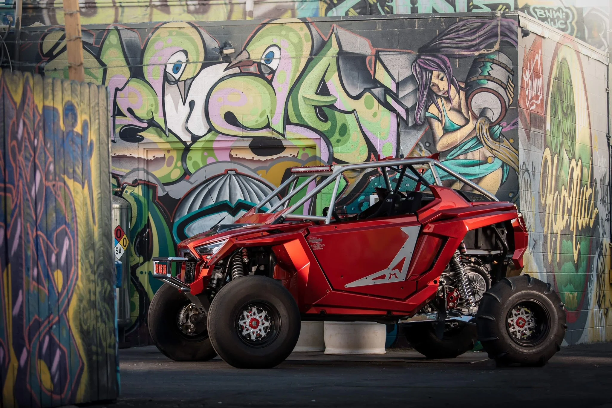 A red off-road vehicle with a metallic roll cage is parked in front of a vibrant graffiti mural on a brick wall. The mural features colorful abstract designs, a cartoonish character, and a woman holding a vinyl record.