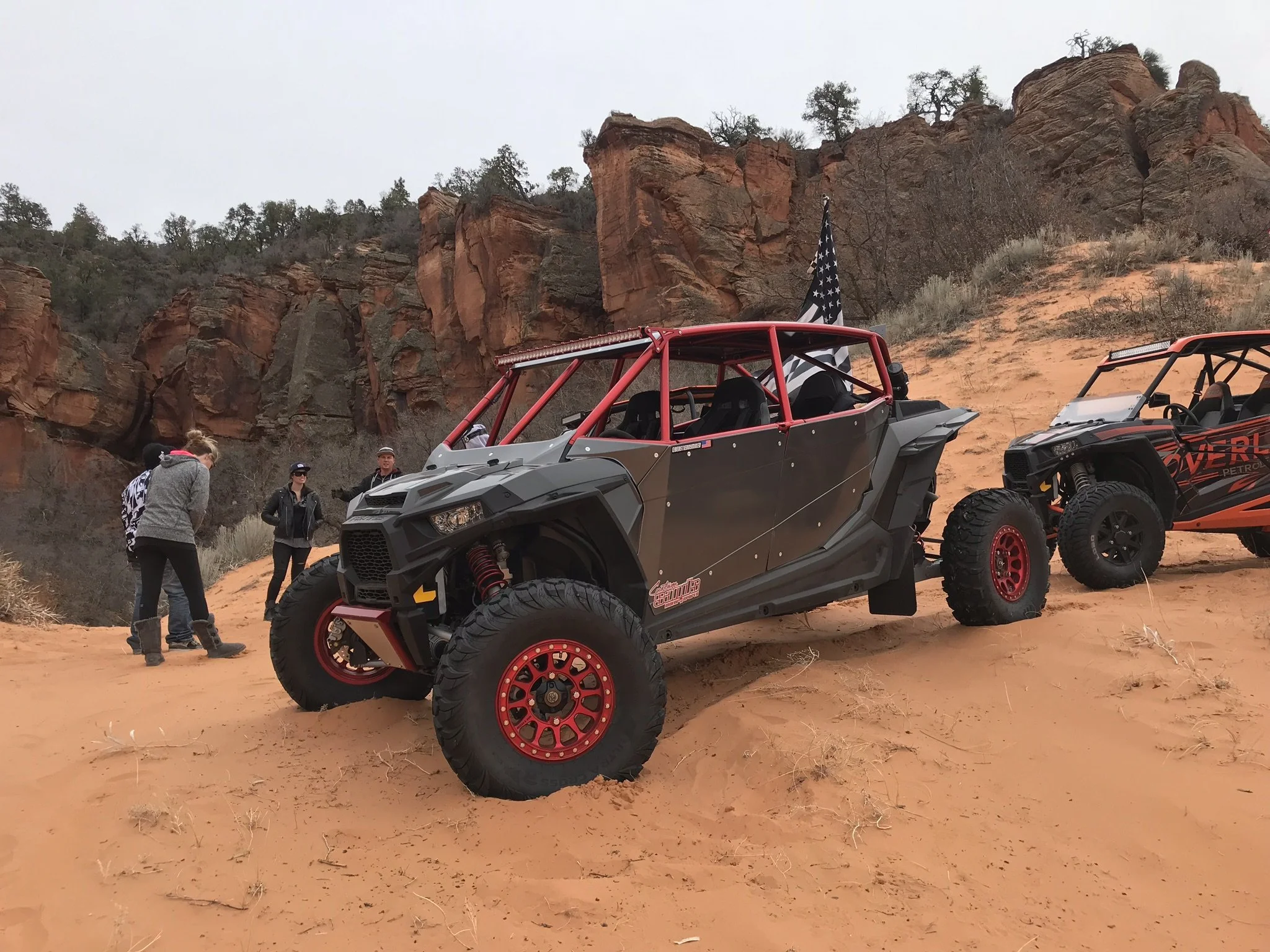 Off-road vehicles parked on sandy terrain with rocky cliffs and sparse vegetation, and three people standing nearby.