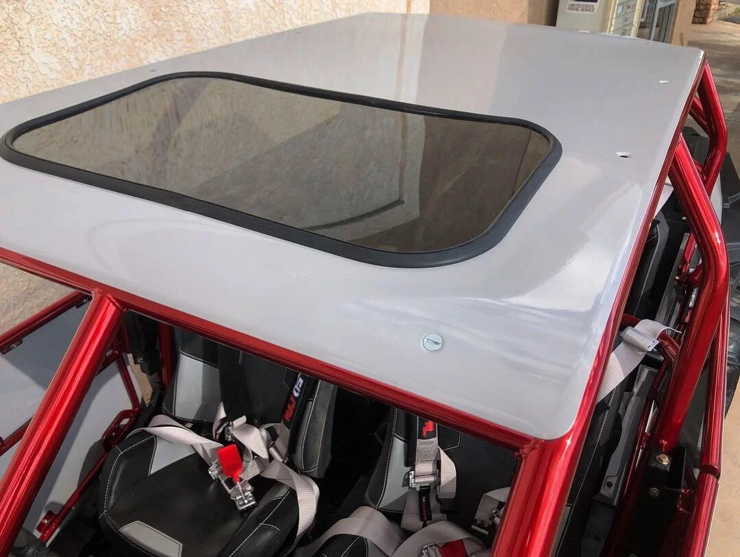 The image shows the interior of a race car, focusing on the black racing seats with harnesses and a silver roof with a tinted sunroof.