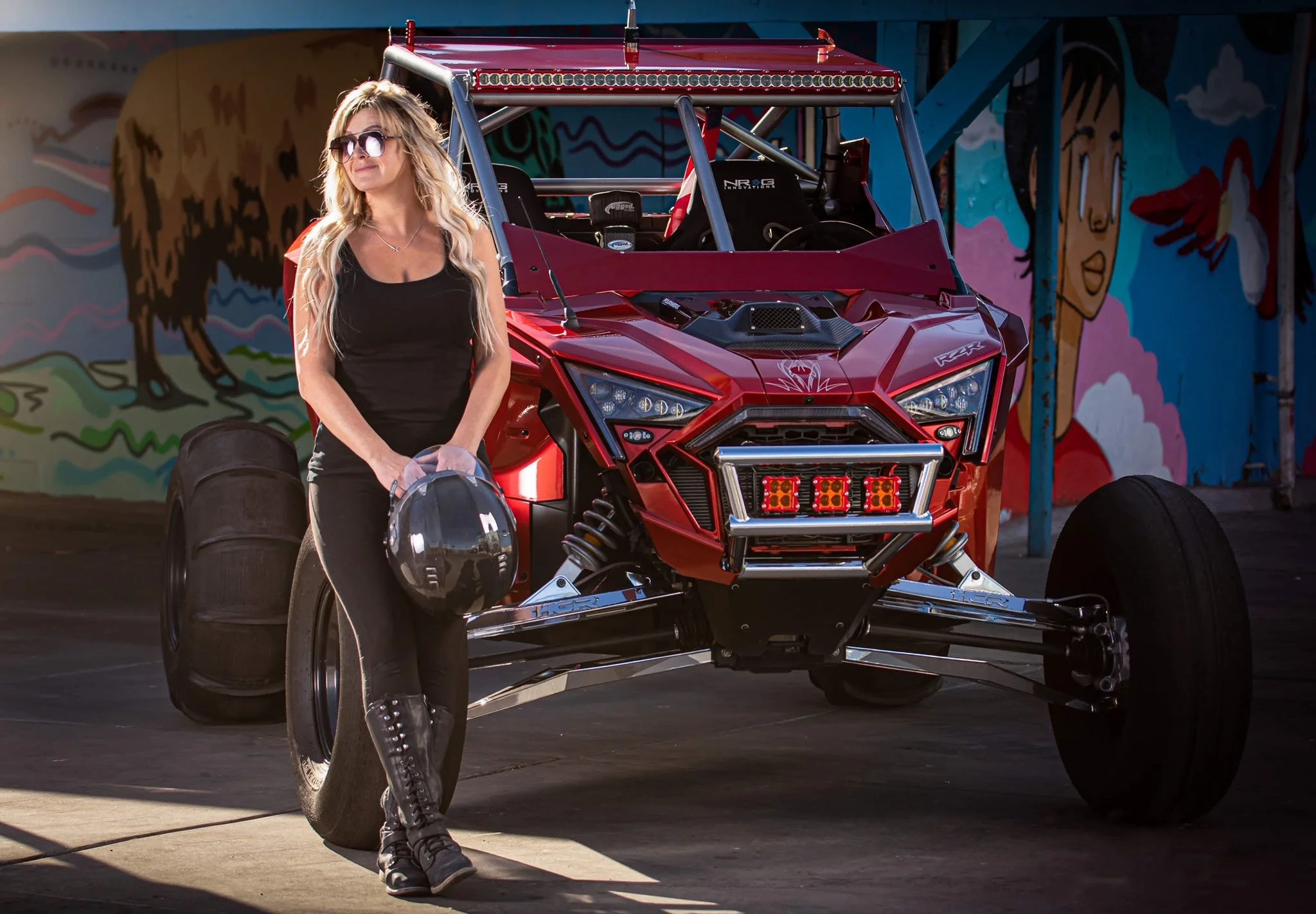 A woman holding a racing helmet and standing next to a red off-road dune buggy with large rear tires, parked in front of a colorful mural.