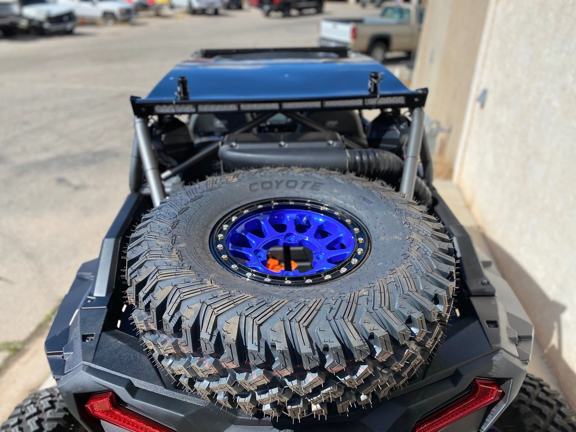 Off-road vehicle with a mounted spare tire on the back, featuring a blue wheel and rugged tread, parked outdoors near a beige wall.