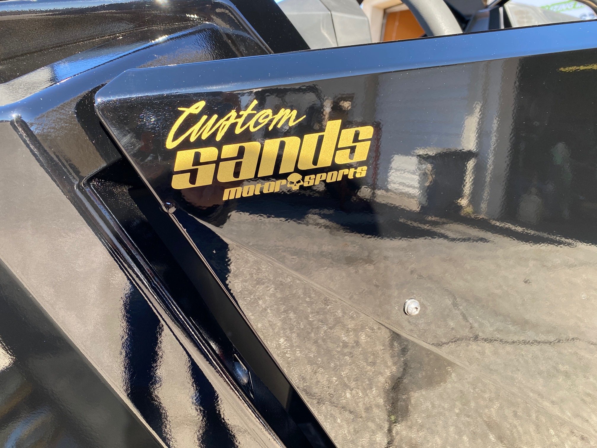 Close-up of a black vehicle with a sticker that says 'Custom Sandi Motor Sports' in gold lettering.