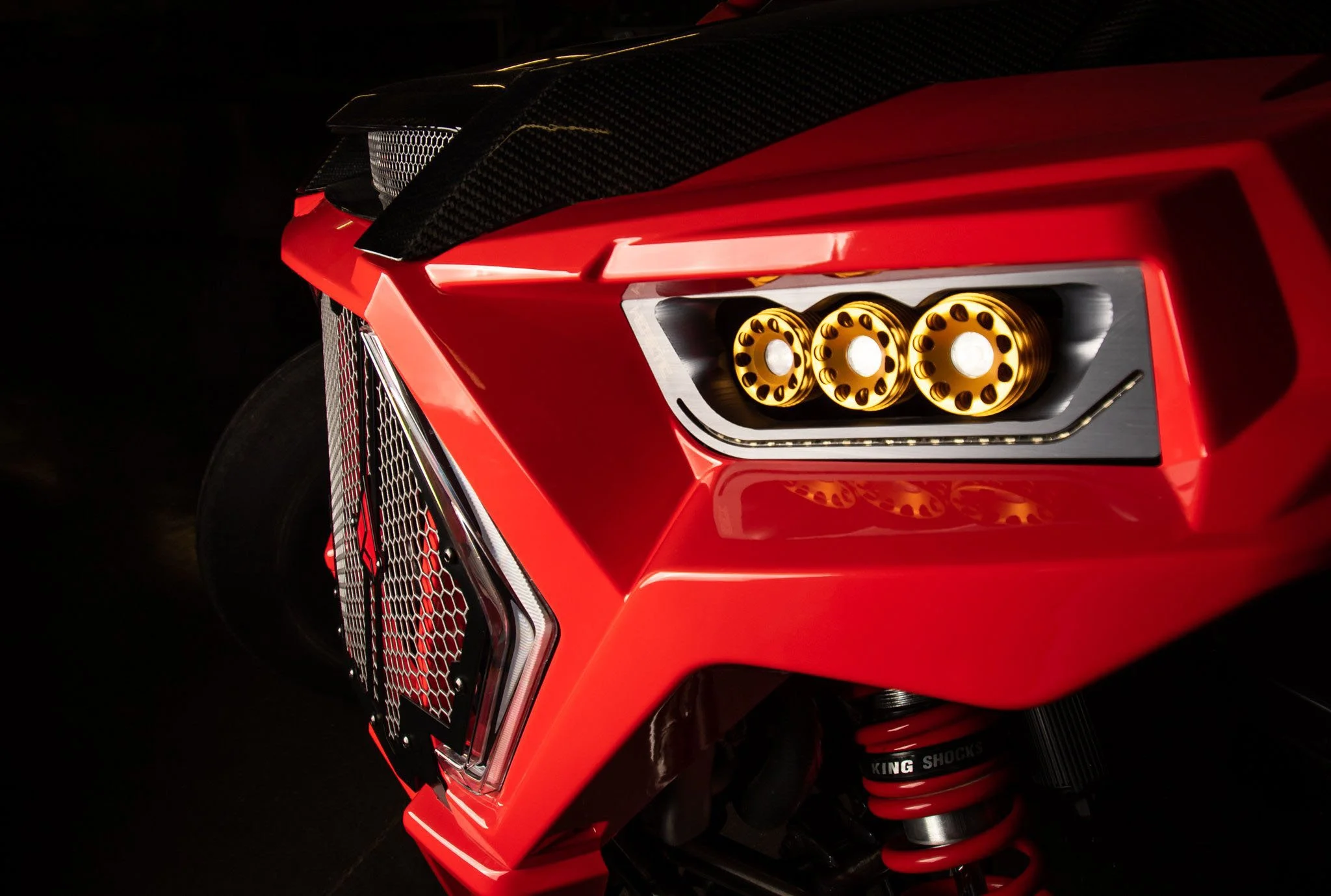Close-up of the front part of a red sports car, focusing on the unique headlight design with three circular golden elements, sleek black detailing, and a front grille with a honeycomb pattern.