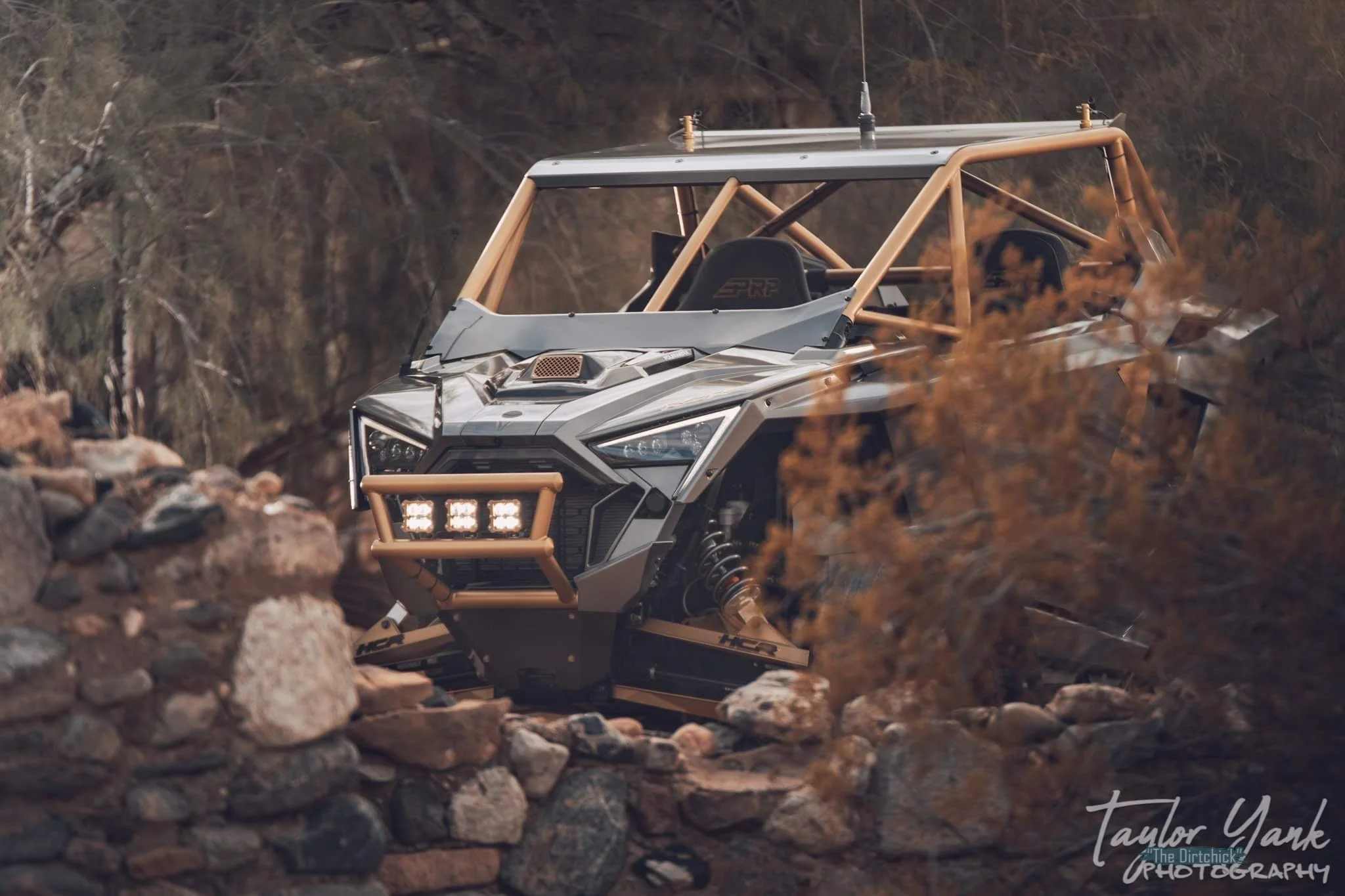 A rugged off-road vehicle driving through a rocky terrain with brownish trees in the background.