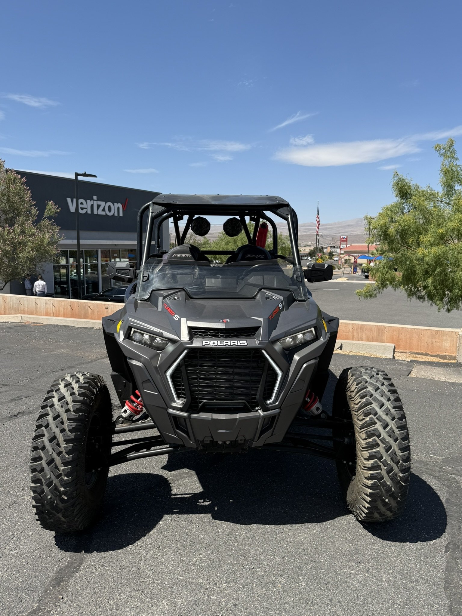 A black off-road dune buggy with large tires, exposed suspension, and a metal frame, parked on a concrete sidewalk in front of a beige wall with two flags on top.