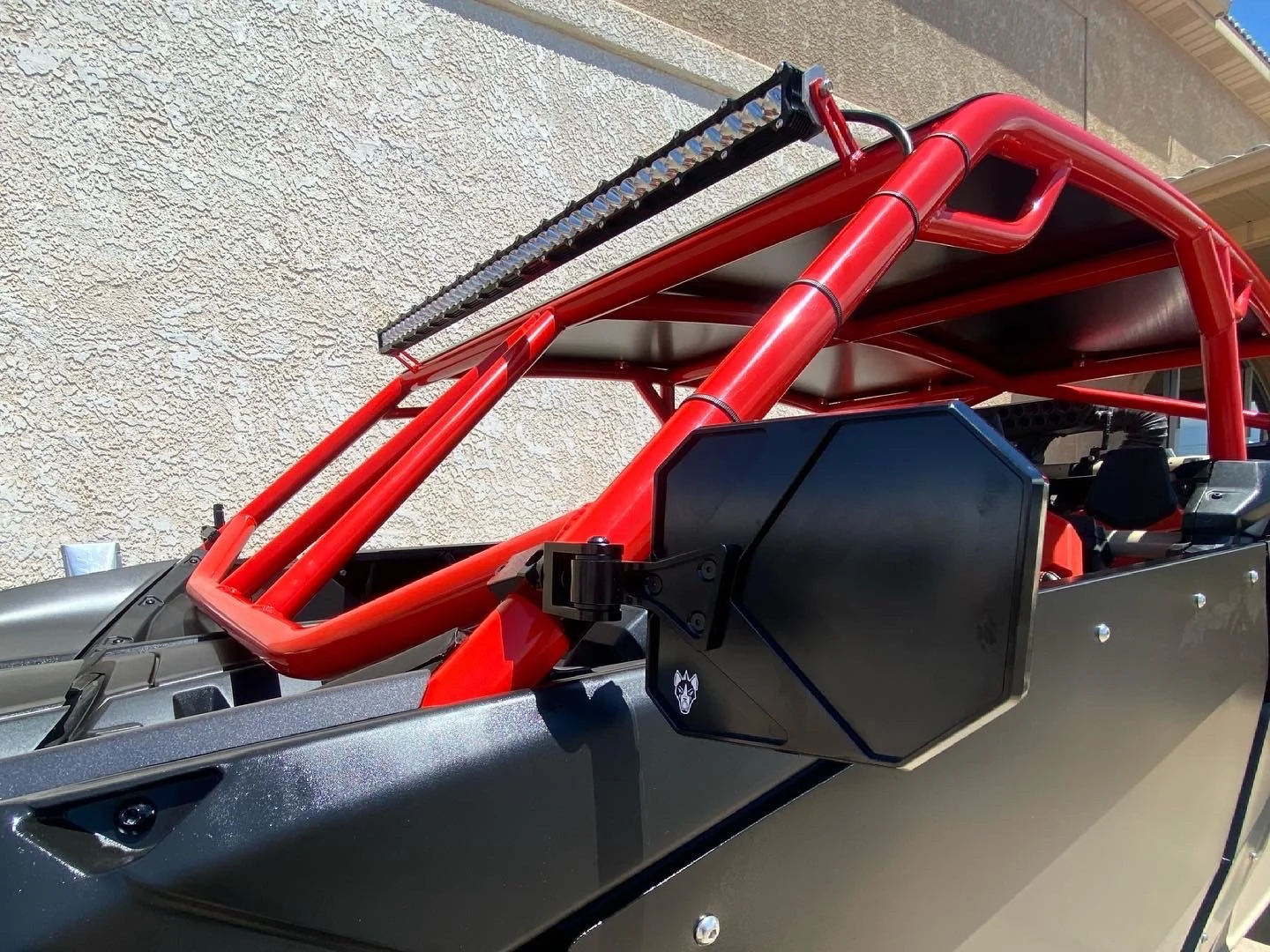Close-up of a race car with a red roll cage, black side panel, and a LED light bar on top, parked beside a beige stucco wall.