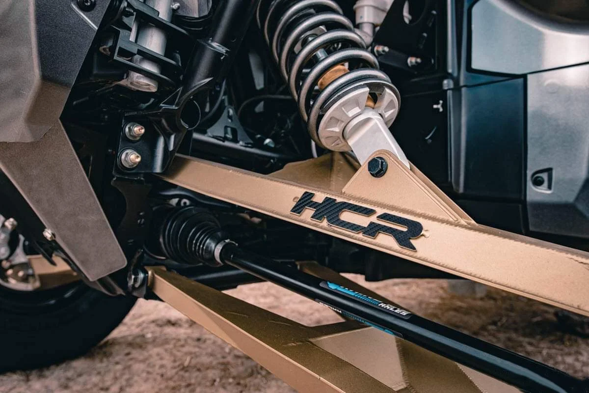 Close-up of a vehicle's suspension system and chassis, showing a beige expediter with the HCR logo, a coil spring, and other mechanical components.