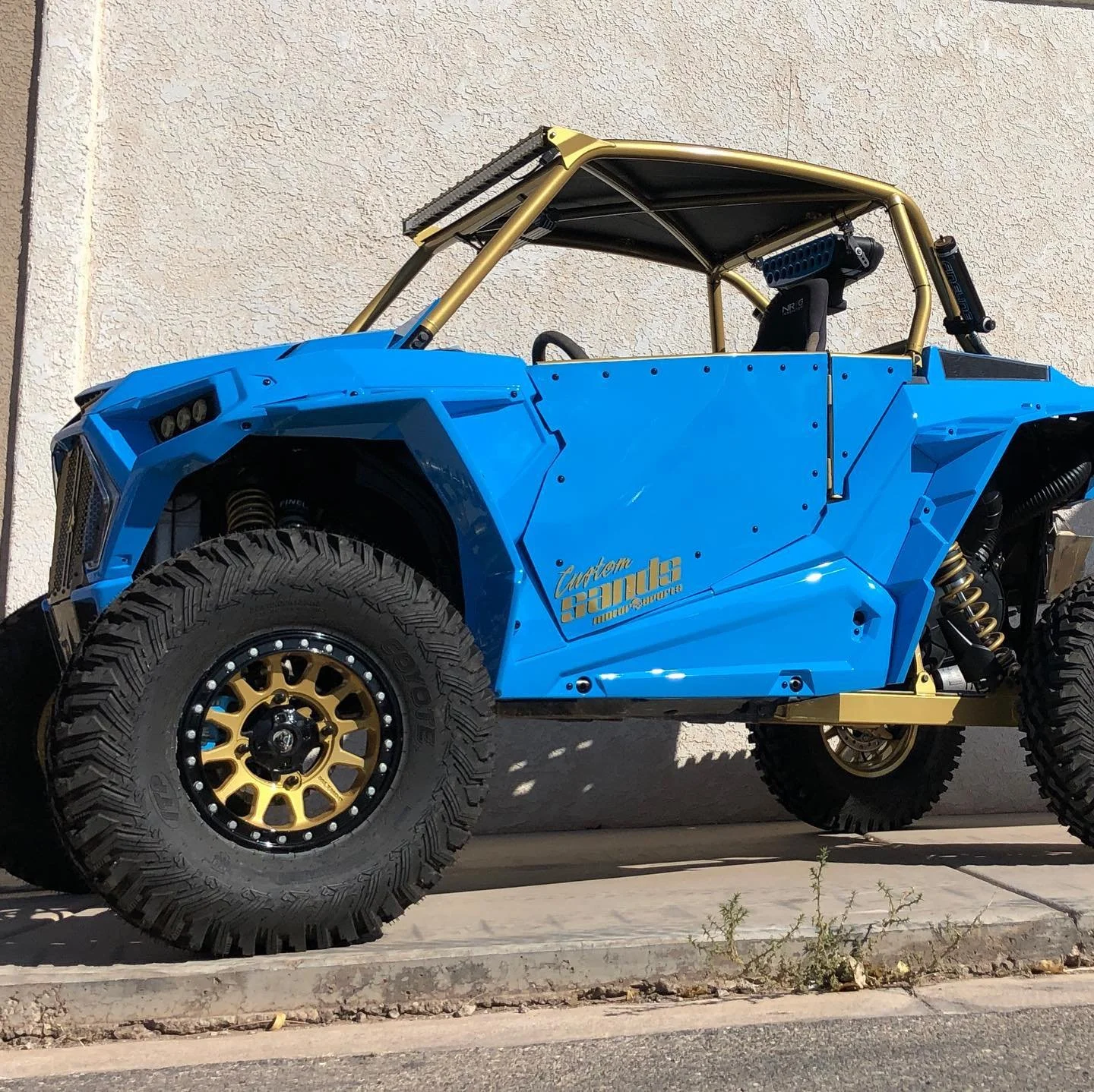 Blue off-road vehicle with large tires, gold wheels, and a gold roll cage parked against a beige wall.