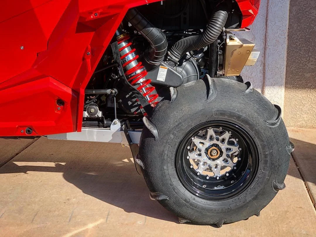 Close-up of a red off-road vehicle's front suspension and tire, showing a large knobby tire and suspension components.