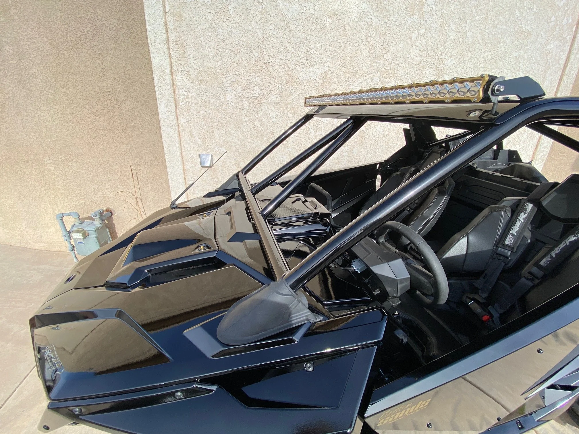 A black off-road vehicle with a roll cage and a light bar on top, parked outside near a beige wall.