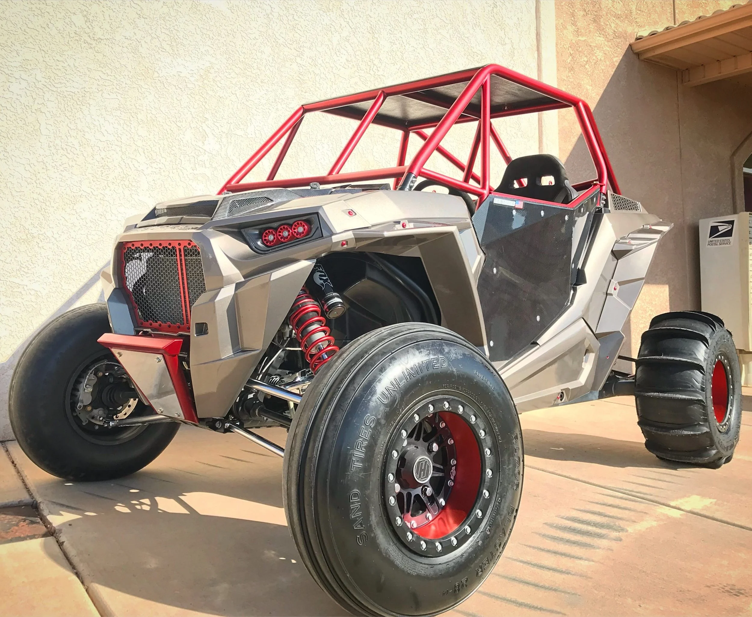 A custom off-road dune buggy with a metallic body, red roll cage, large off-road tires, and visible suspension system, parked on a concrete surface near a beige wall.