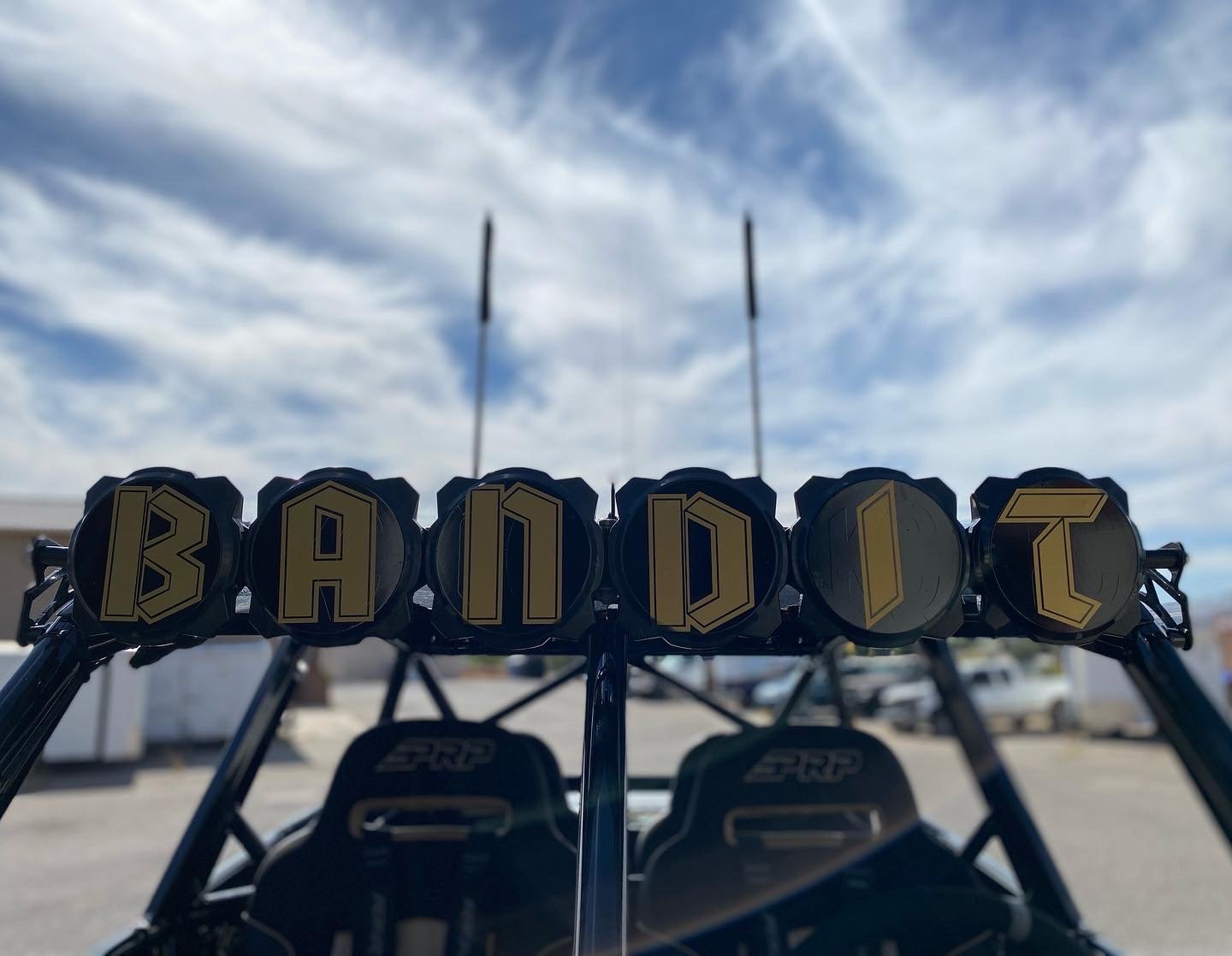 A row of golf clubs on a golf cart with black seats, with the word 'BADK1T' spelled out on the clubs, outdoors under a partly cloudy sky.