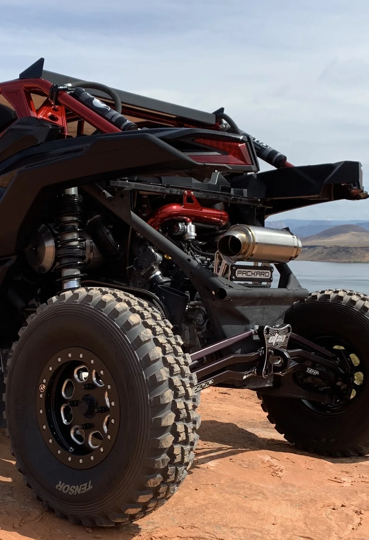 Off-road vehicle with large tires and exposed mechanical components, set against a landscape with water and mountains.
