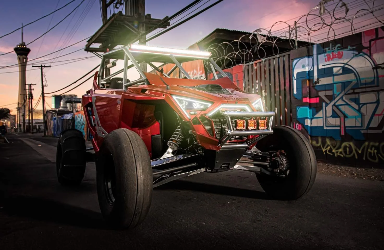 A custom off-road vehicle with a red body, exposed mechanical parts, and large tires is parked on a street during sunset. The vehicle has a roll cage and LED lights on the front.