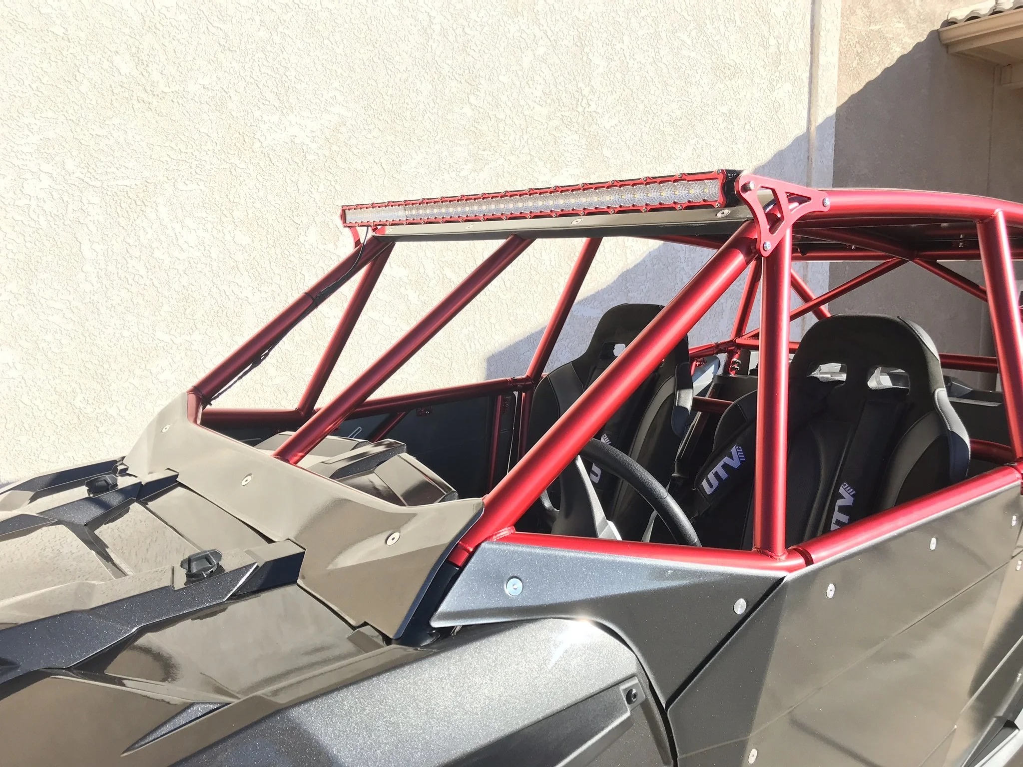 A metallic off-road racing vehicle with a red roll cage and seats, parked against a plain wall.