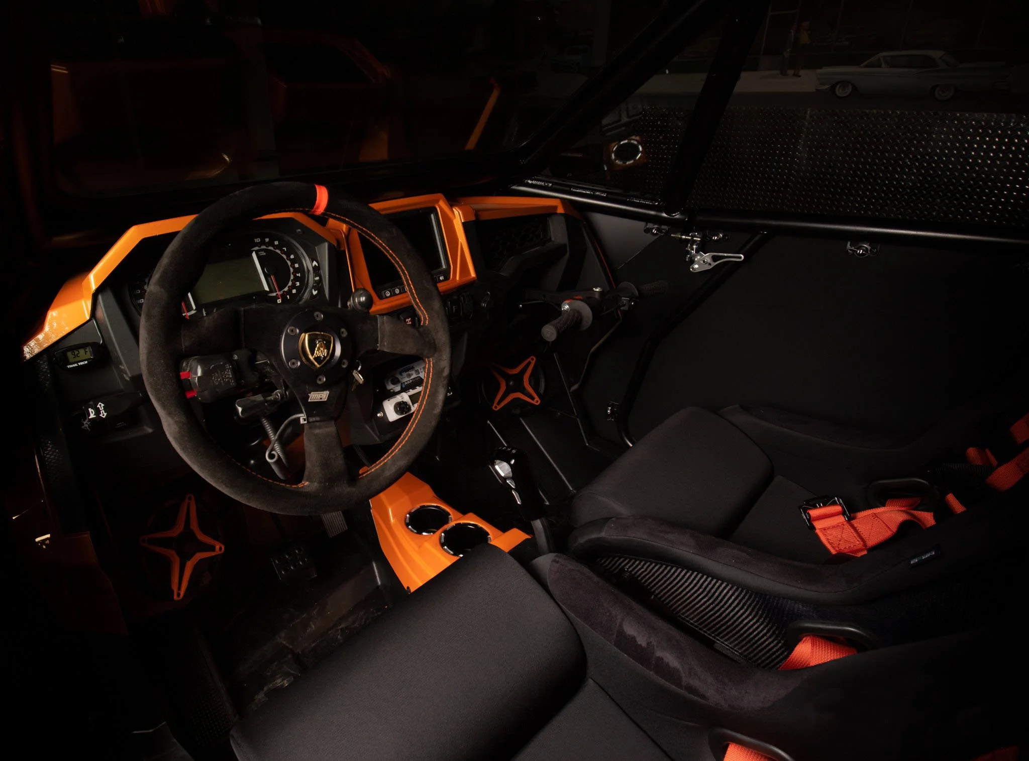 Interior of a Lamborghini race car with black and orange accents, racing seat with orange straps, and a steering wheel with Lamborghini logo.