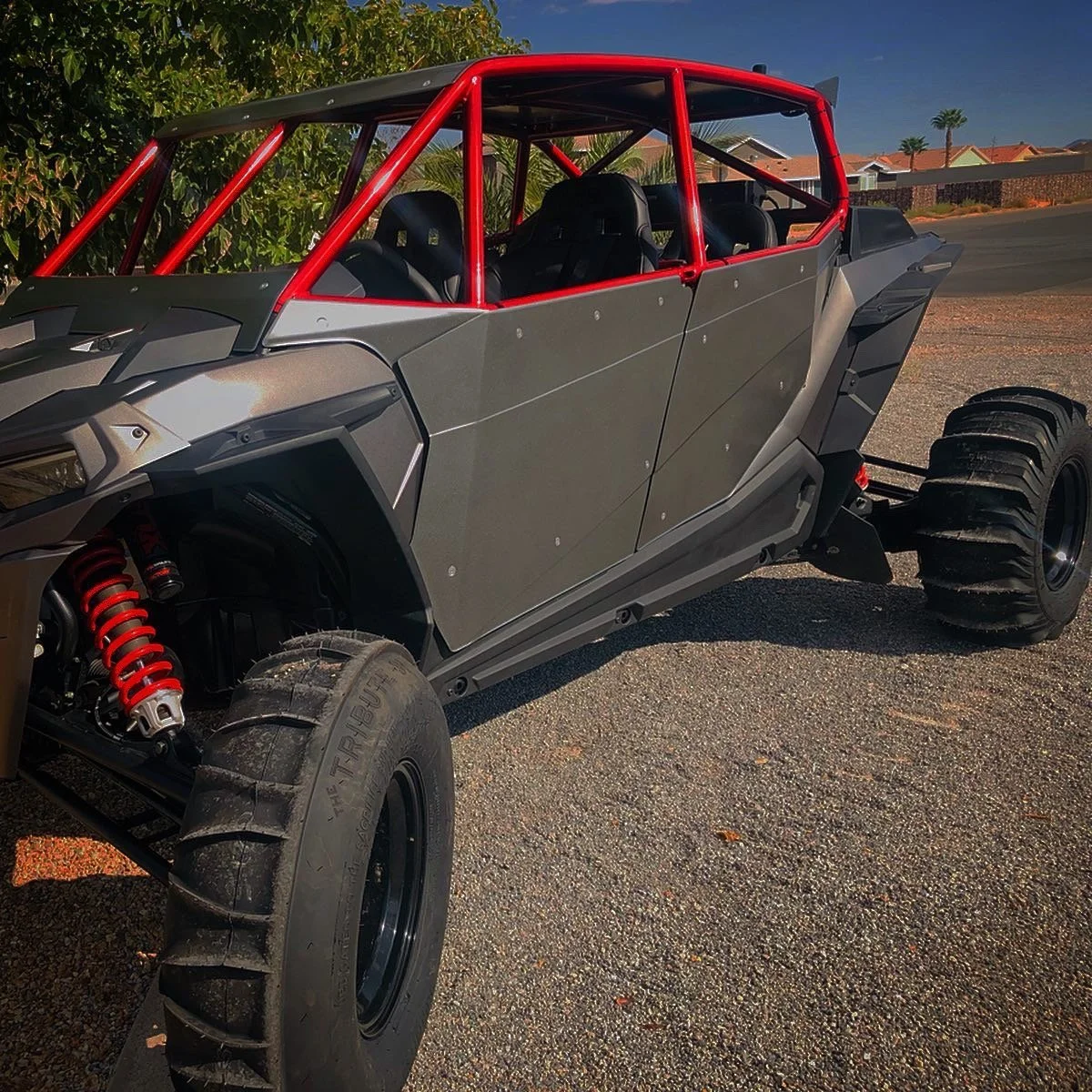 A custom-built off-road vehicle with large tires, red suspension shocks, gray body panels, and a red roll cage, parked on a paved surface with residential houses and palm trees in the background.