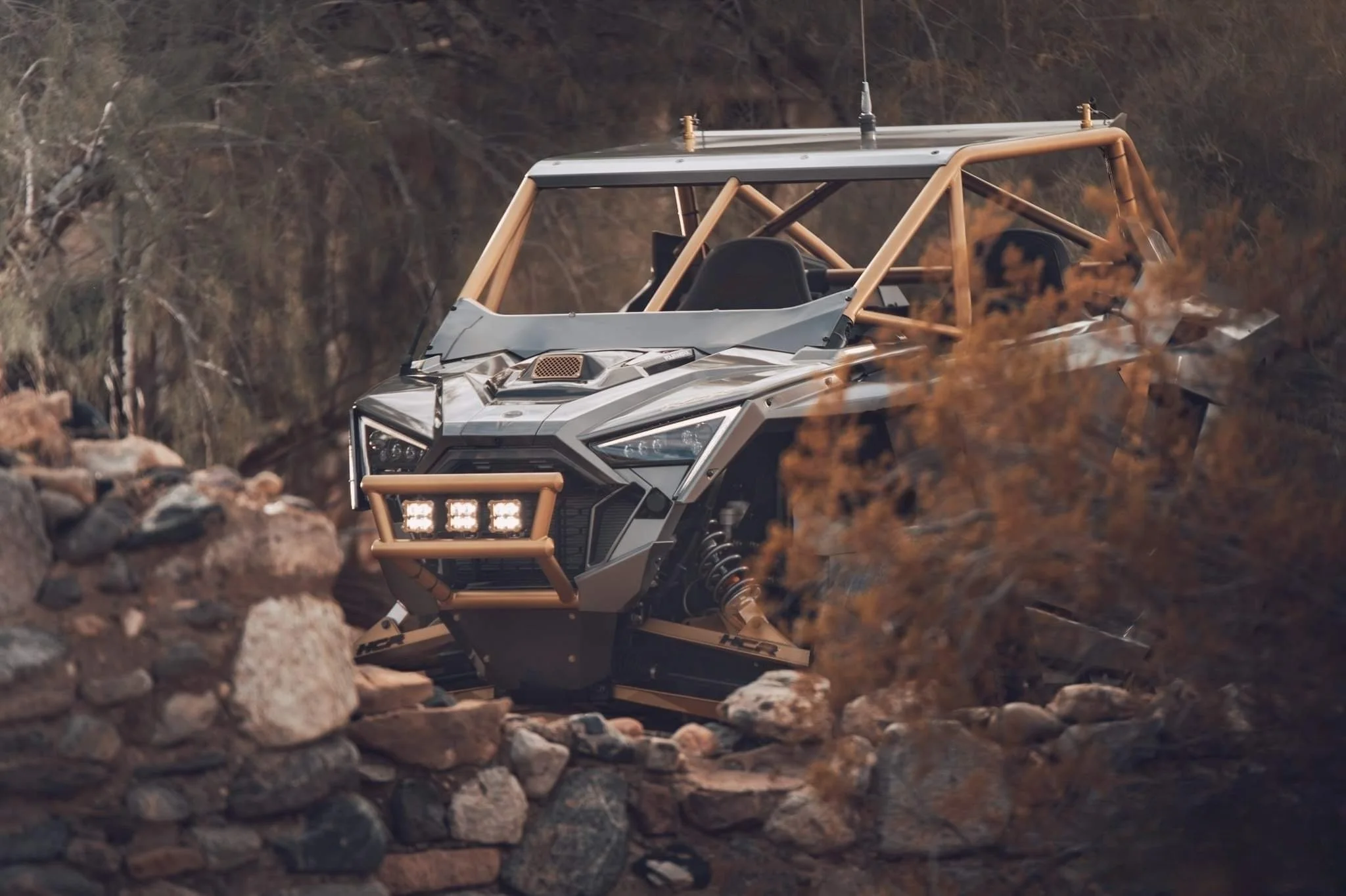 An off-road vehicle with a metallic body and a tan roll cage navigating rocky terrain with autumn foliage in the background.