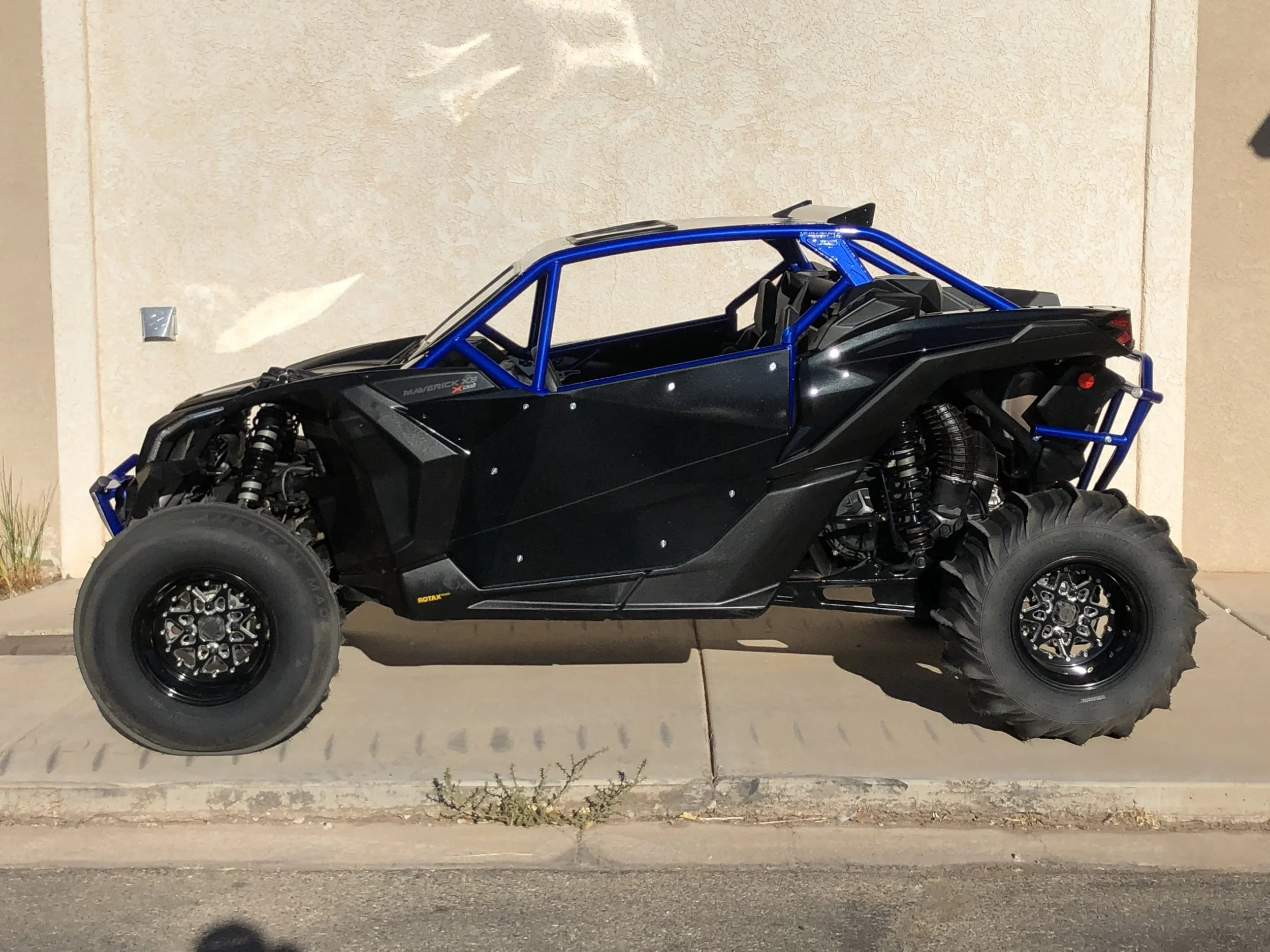 Black off-road vehicle with blue roll cage parked on sidewalk in front of beige wall.