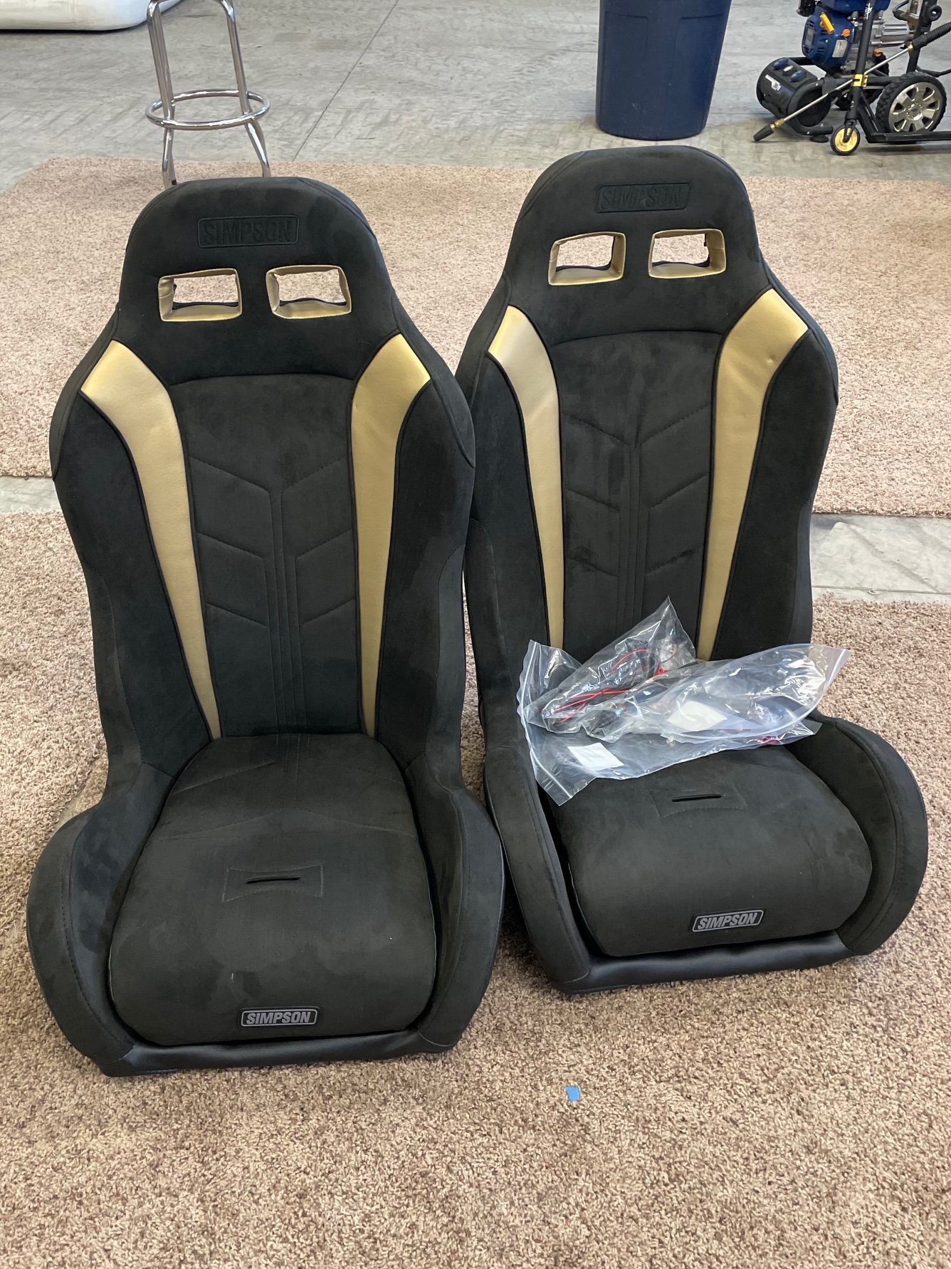 A pair of black and yellow Simpson racing seats placed on a beige carpet in an indoor setting with various objects in the background.