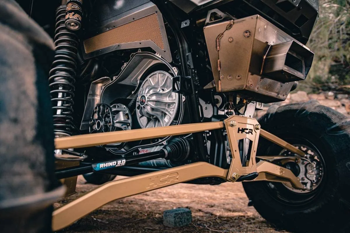 Close-up view of a rugged off-road electric vehicle's suspension and drivetrain components, including a gold-colored suspension arm, motor, and large tires, set outdoors on dirt.