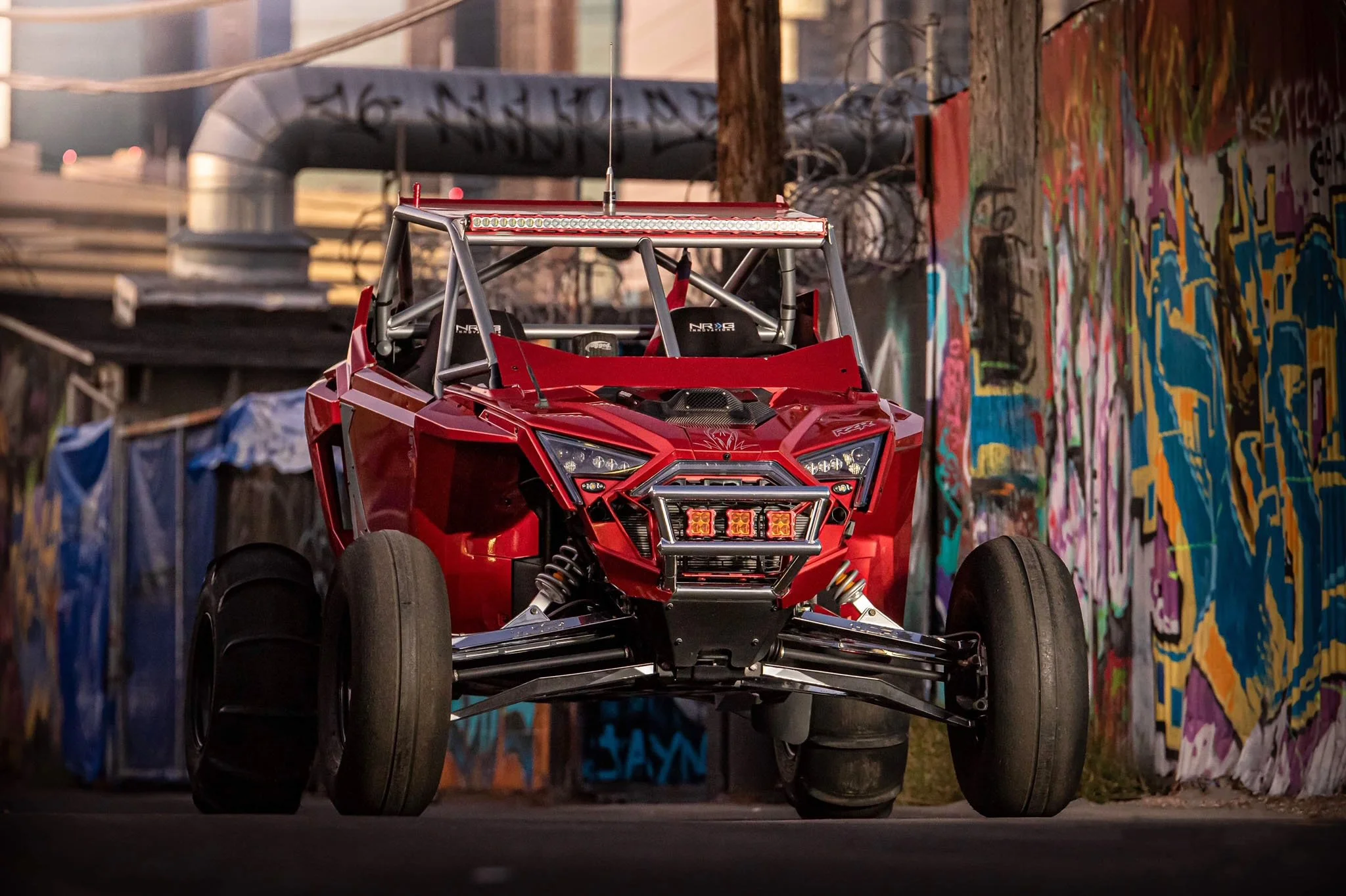 Red off-road racing vehicle with exposed suspension and LED light bar, parked against graffiti-covered walls and industrial background.