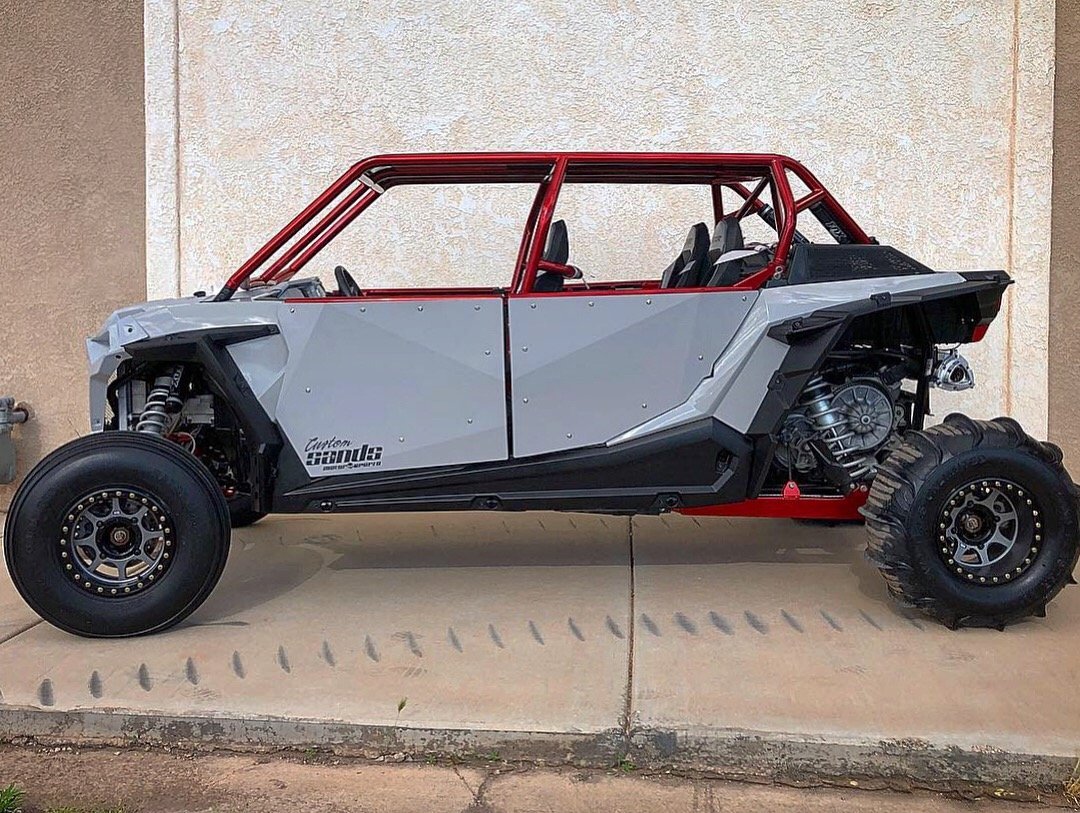 A custom off-road vehicle with a partially assembled frame, large tires, and exposed engine components, parked on concrete.