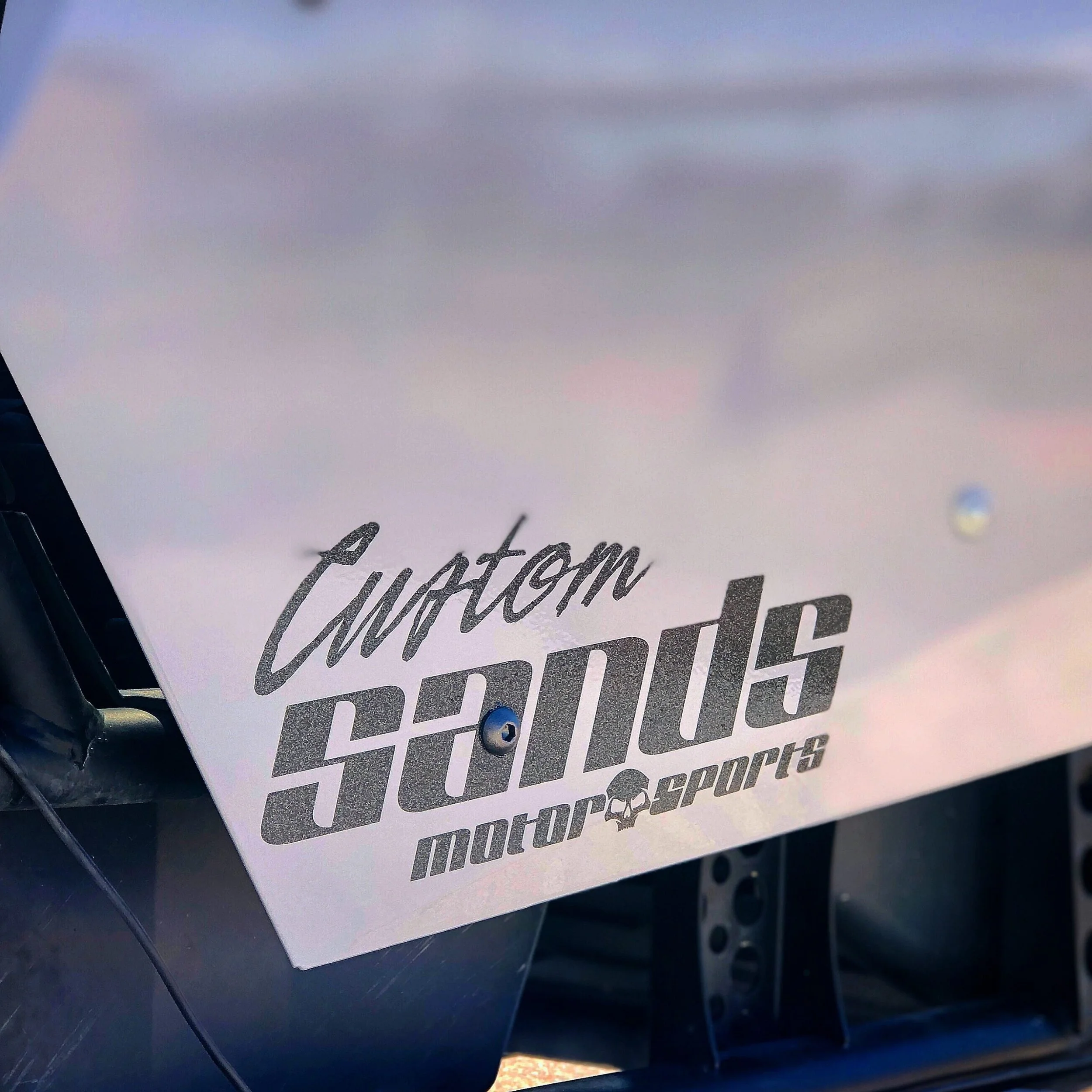 Close-up of a white sign with the words 'Custom Stands Motor Sports' printed on it, attached to a vehicle or equipment with visible screws.