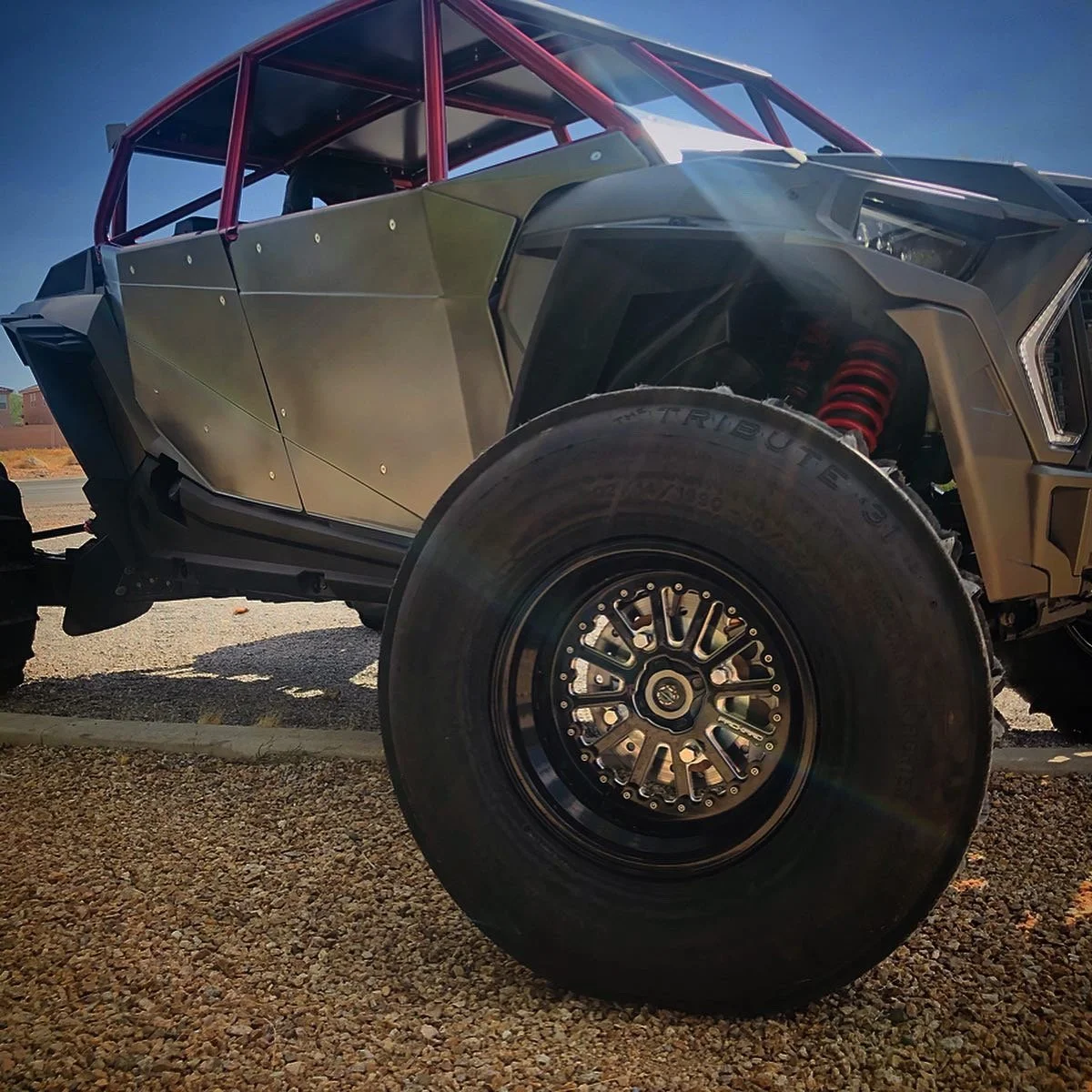 Close-up of a futuristic off-road vehicle with large tire and exposed suspension, parked outdoors on gravel with a clear blue sky.