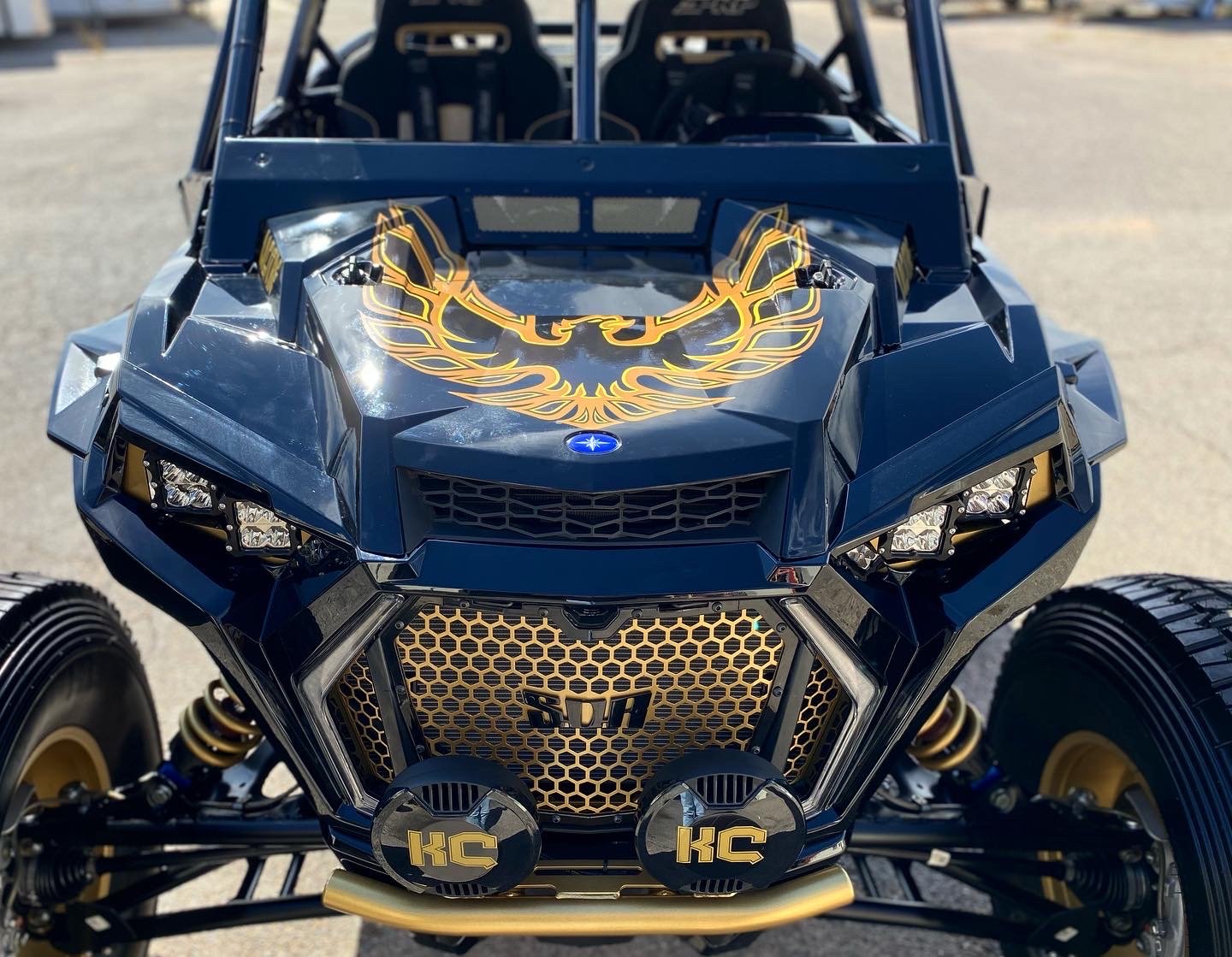 A heavily modified Polaris Slingshot three-wheeled vehicle with black and gold accents, featuring a phoenix decal on the hood, KC off-road lights, and racing seats.