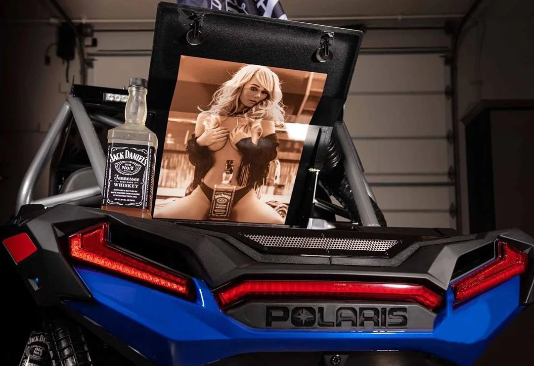 A toy Polaris vehicle with a framed photo of a woman, a Jack Daniel's whiskey bottle, and a small whiskey bottle inside it, placed in a garage.