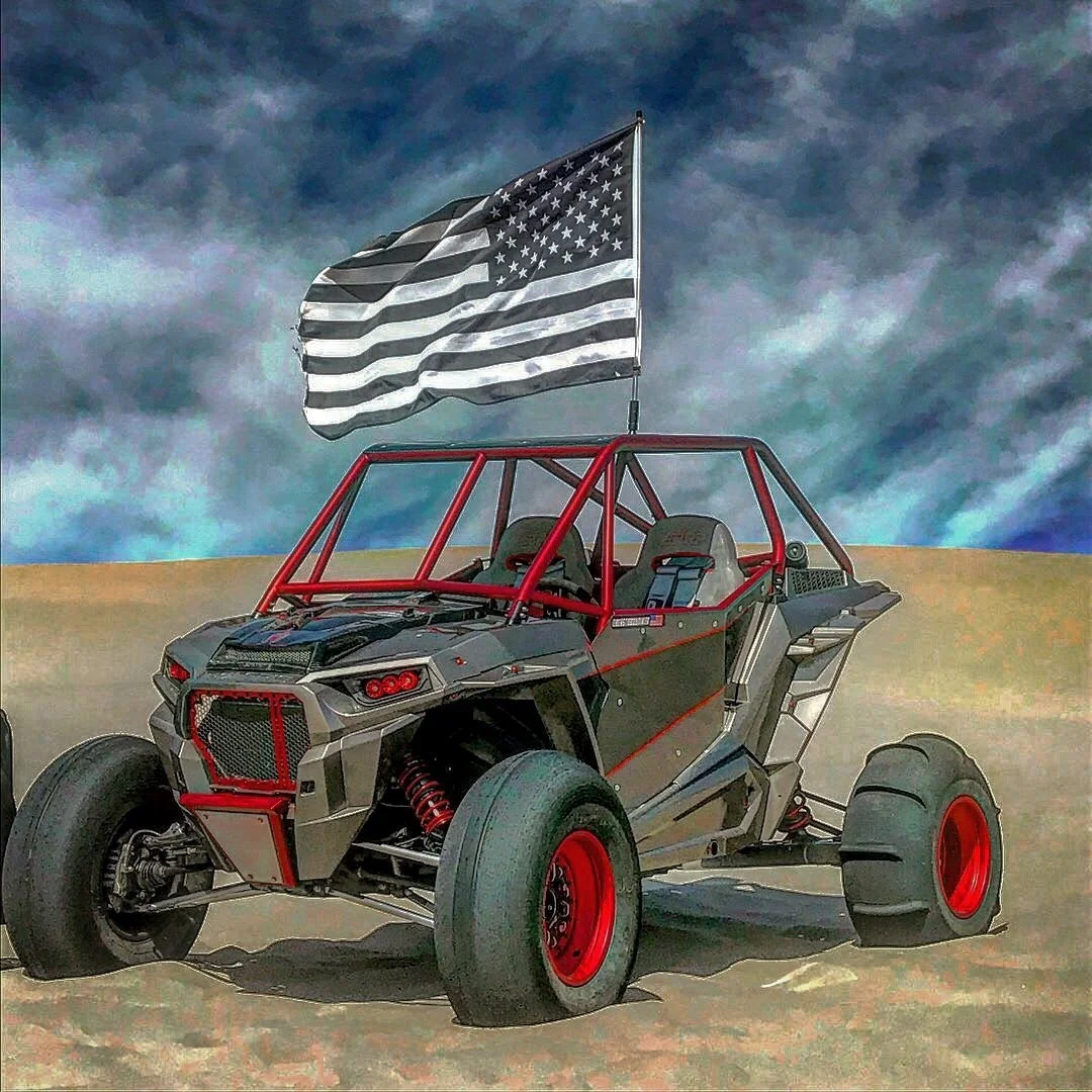 An off-road vehicle with a black and gray body, red frame and wheel rims, and large tires, flying a black and white American flag on a pole, on a sandy terrain under a cloudy sky.