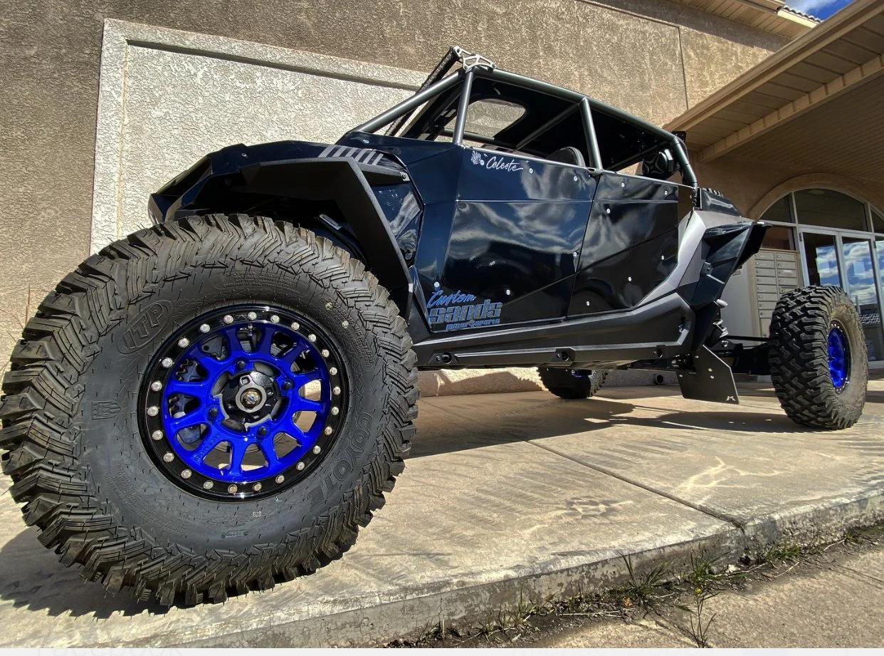 A black off-road vehicle with large tires and blue rims parked on a concrete driveway near a beige house with arched windows.