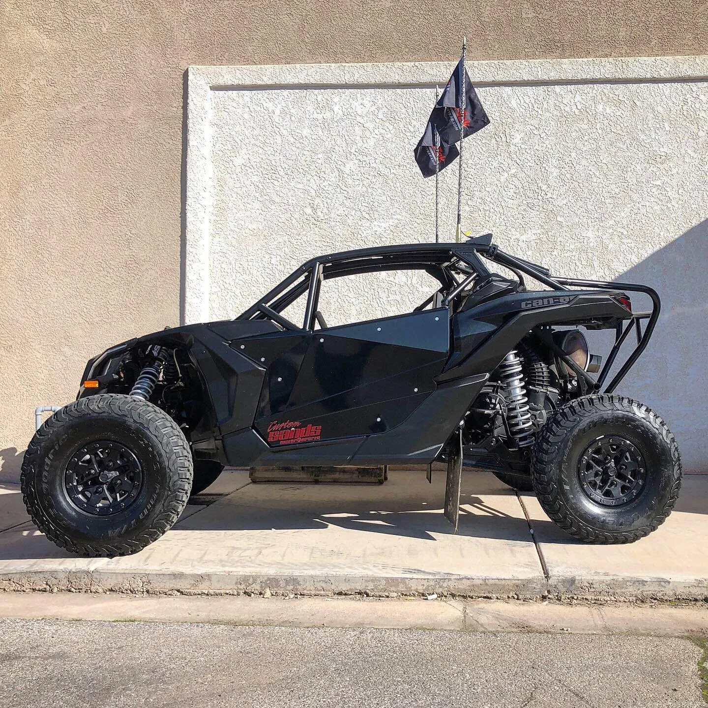 A black off-road dune buggy with large tires, exposed suspension, and a metal frame, parked on a concrete sidewalk in front of a beige wall with two flags on top.