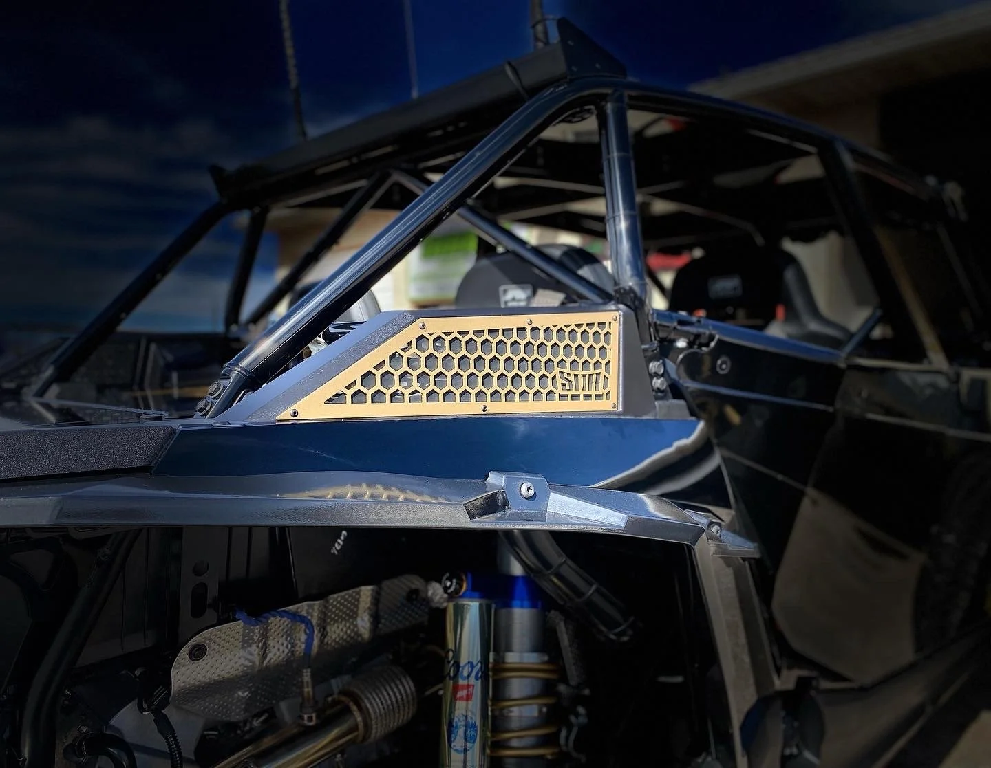 Close-up view of a race car's interior and engine components, featuring a roll cage, a gold-colored honeycomb air vent, and visible suspension parts.