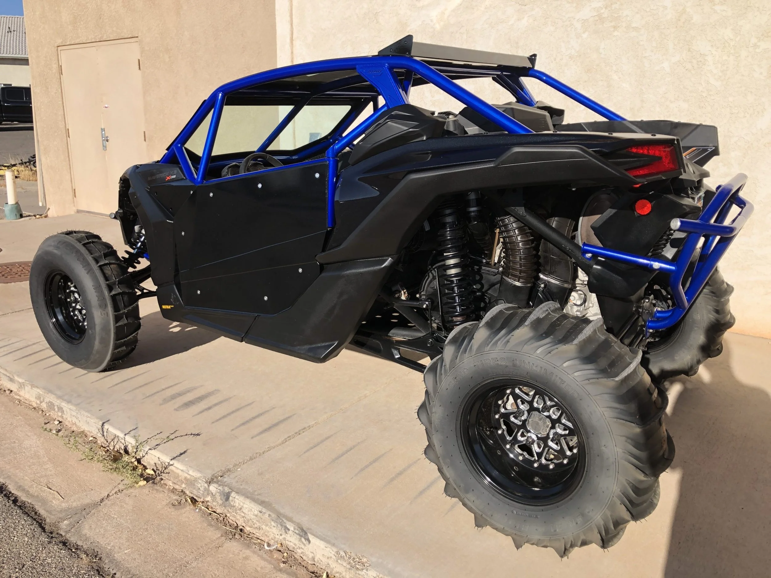 Off-road vehicle with large tires, black body, blue roll cage, and exposed suspension parts parked on a concrete sidewalk.