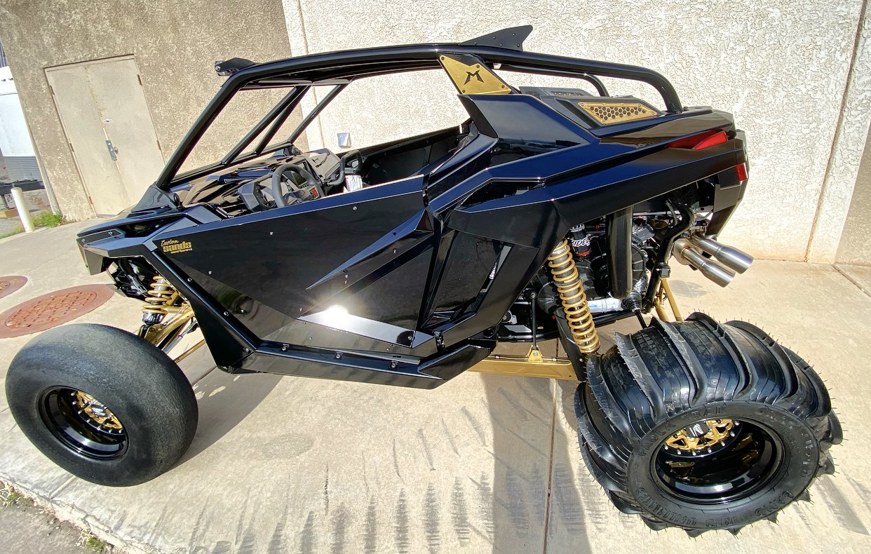 A custom-built, black and gold off-road dune buggy with large tires, suspension, and a sleek, angular body design parked outdoors on concrete.