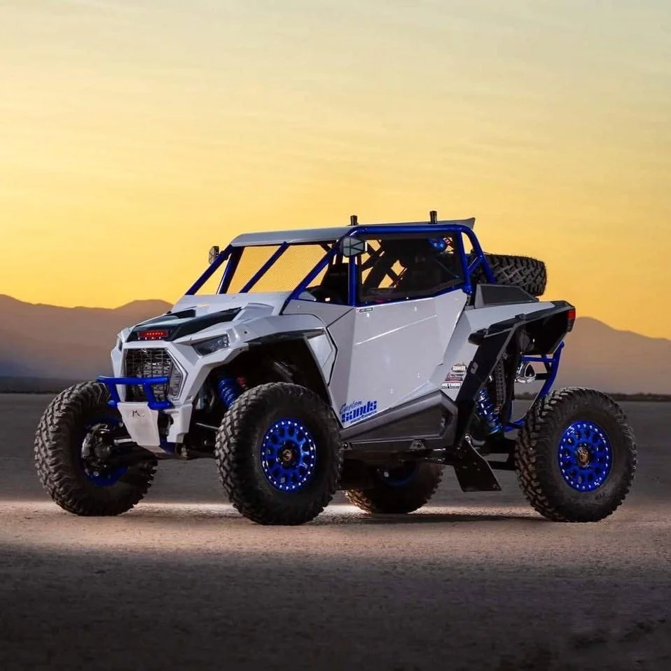Off-road racing vehicle on desert terrain at sunset with distant mountains, white body, blue accents, large tires, and spare tire on back.
