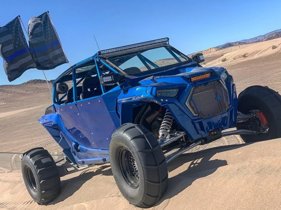A blue off-road vehicle with large tires, flag decals, and a light bar on top, parked on a desert landscape with sand dunes and mountains in the background.