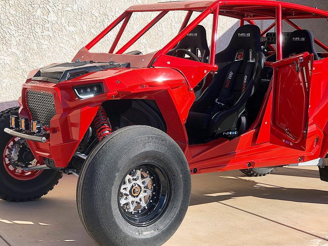 A red off-road vehicle with a roll cage, racing seats, and large tires, parked against a beige wall.