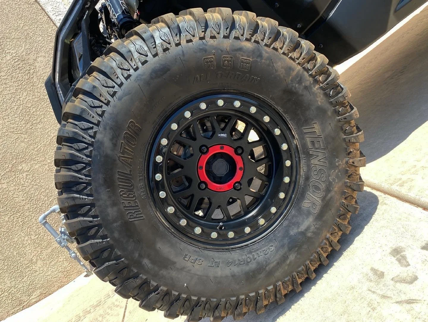 A close-up of a large off-road tire mounted on a black rim with red accents, branded TENSER and REBOUR TOURE, with a textured beige wall and concrete ground in the background.