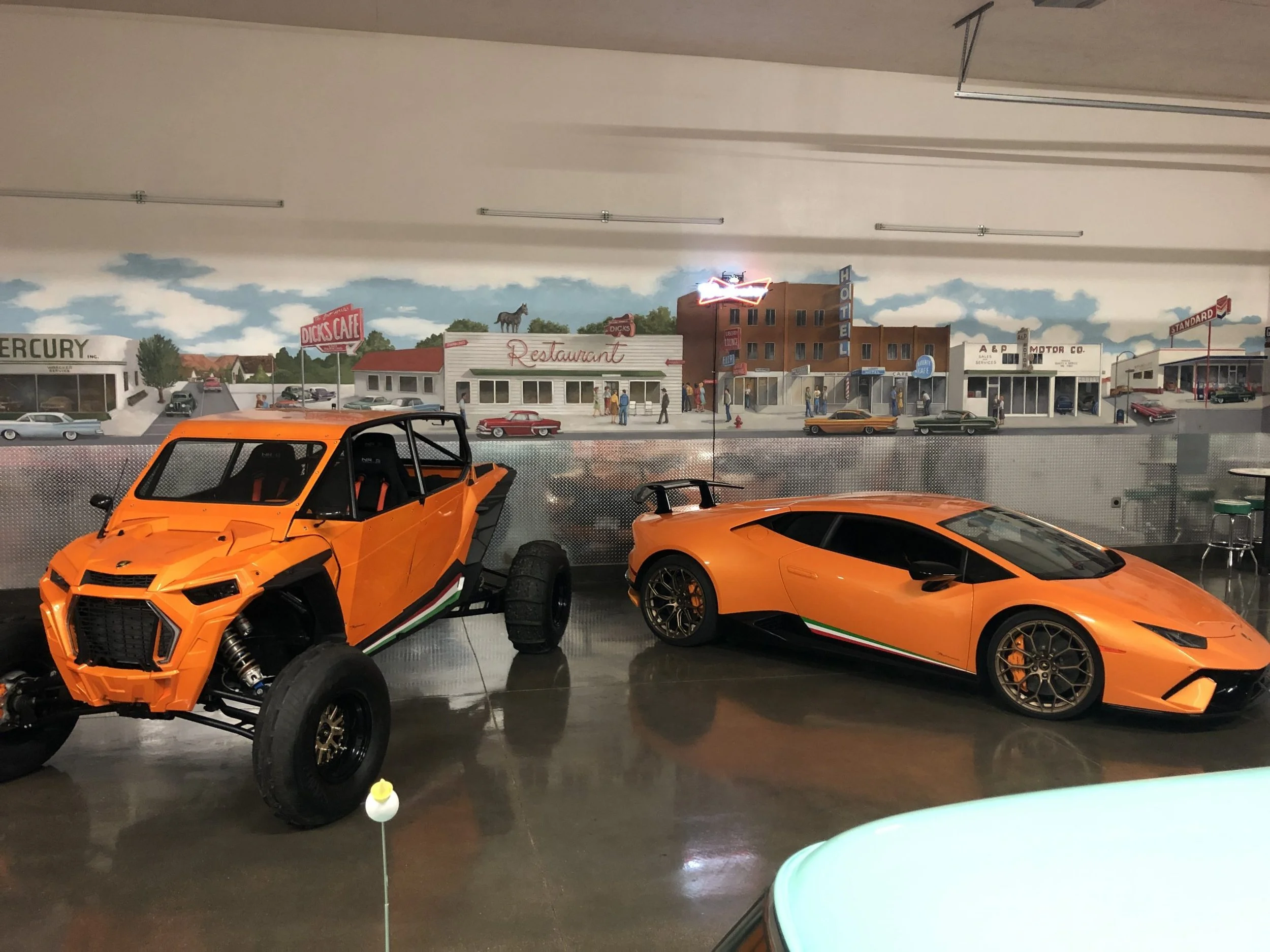 Orange off-road vehicle and a Lamborghini parked inside a museum with a retro city mural in the background.