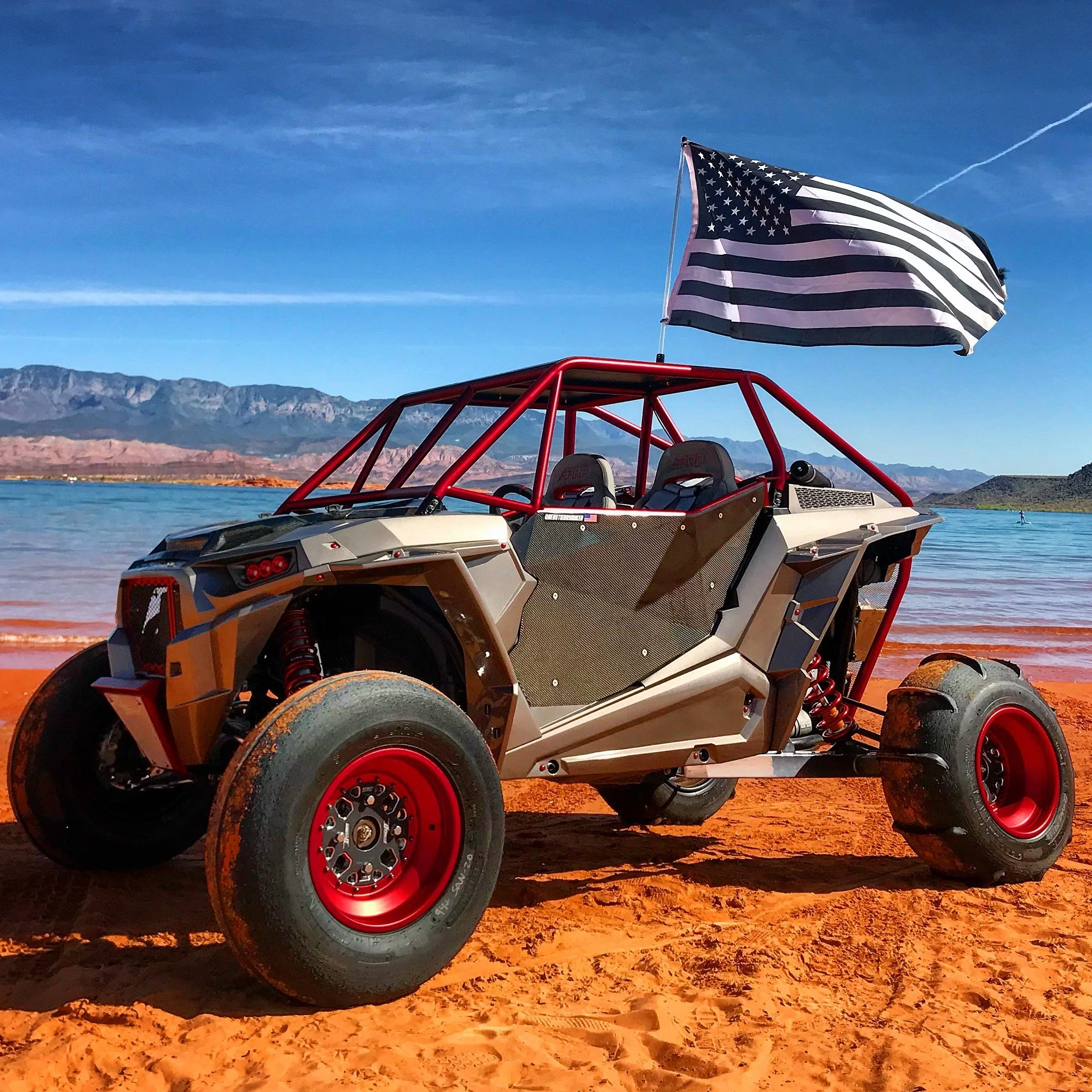 Off-road vehicle parked on a sandy beach with an American flag attached, set against a lake and mountain landscape under a blue sky.
