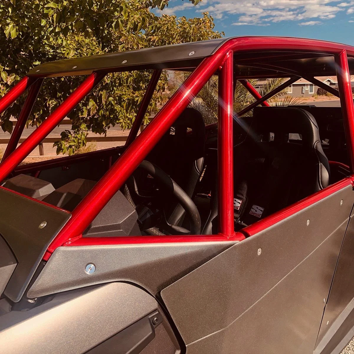 Interior of a race car with a red roll cage, black racing seats, and a dashboard, parked outdoors with trees and a clear sky in the background.