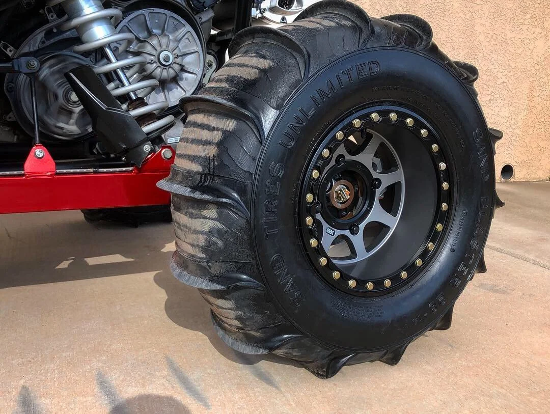 Close-up of a large off-road tire with the words 'SAND TRIPS UNLIMITED' written on it, mounted on a black rim with yellow lug nuts. Part of a vehicle's suspension system and engine are visible in the background.