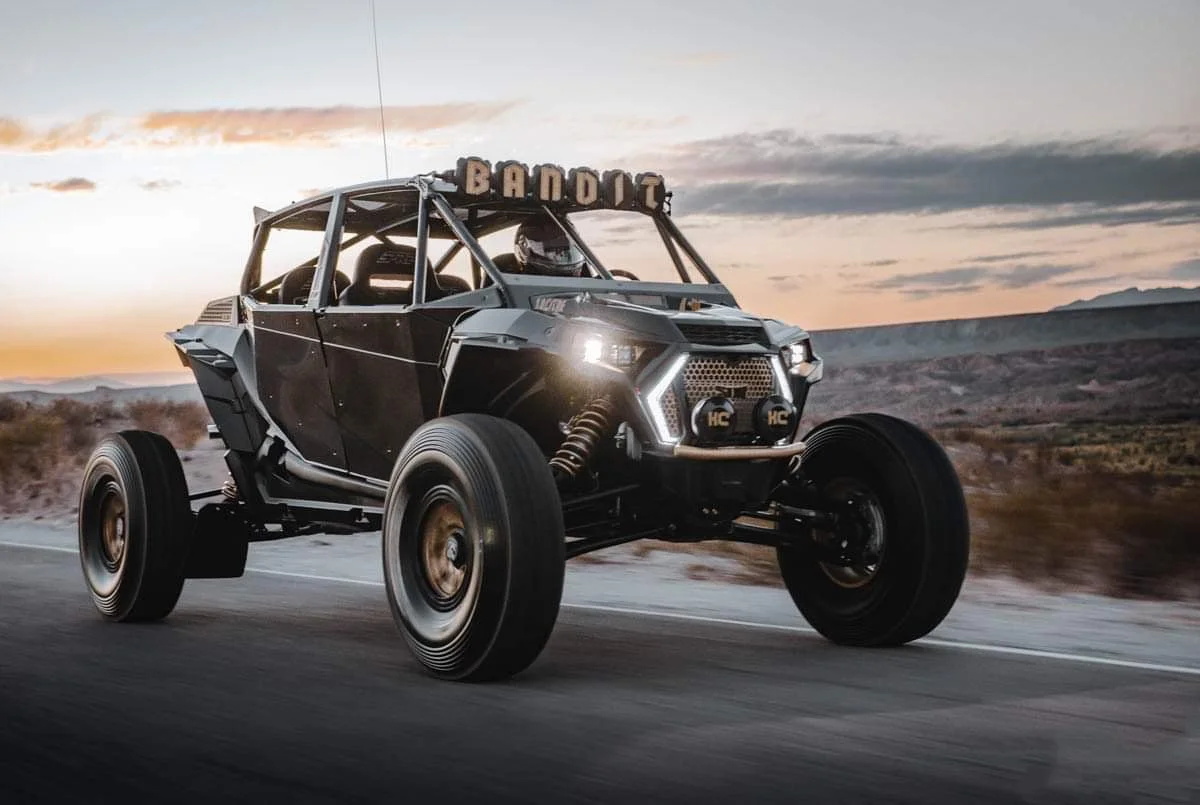 An off-road dune buggy driving on a dirt road during sunset in a desert landscape.