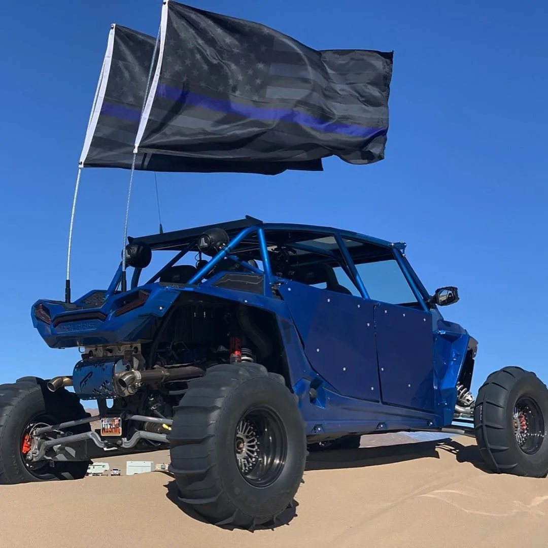 A blue off-road vehicle with large tires and a black American flag with a blue stripe waving in the wind, set against a clear blue sky.