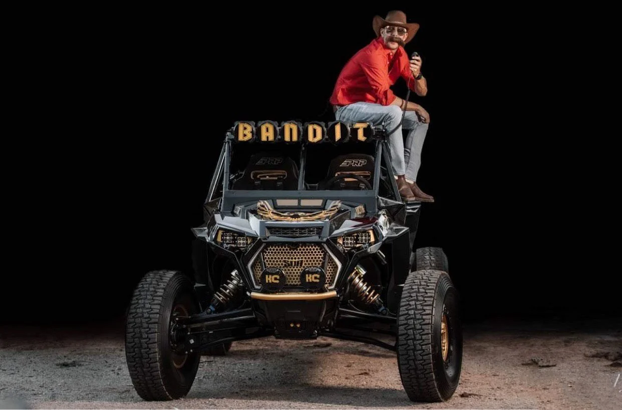 A man wearing a cowboy hat, sunglasses, and a red shirt sits on top of a black off-road vehicle with large tires. The vehicle has a sign that says 'BANDIT' on the roof and features gold accents and high-performance suspension.