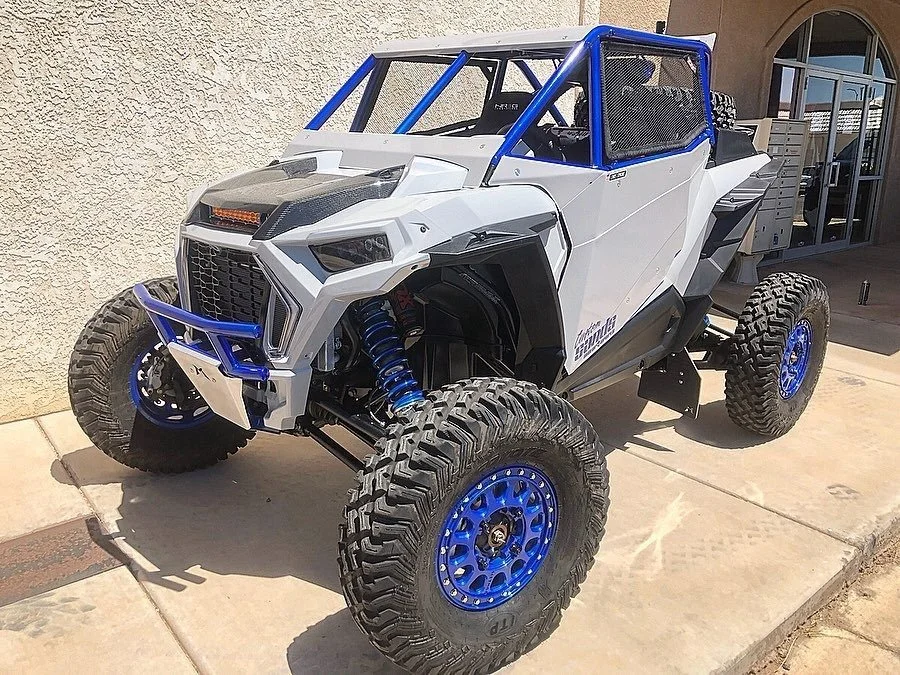 Off-road buggy with large tires, white and blue body, and protective roll cage, parked on sidewalk in front of a building.