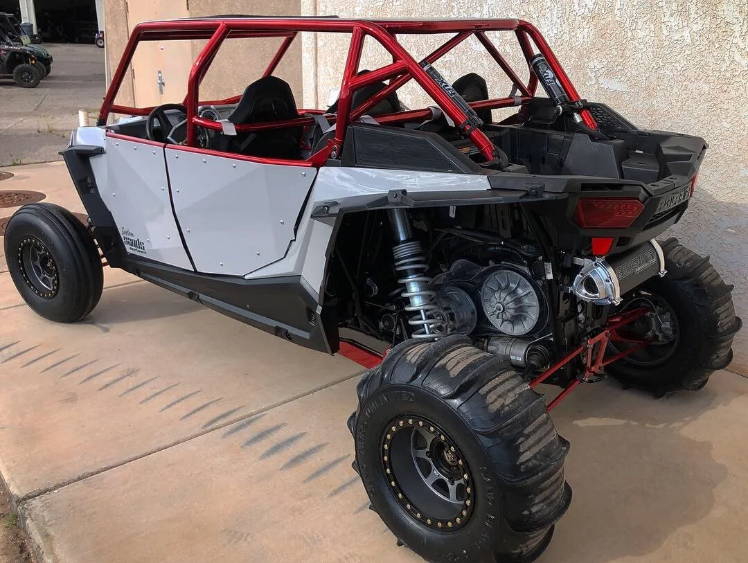 A custom off-road vehicle with a roll cage, exposed mechanical components, and large rugged tires, parked on a concrete driveway.